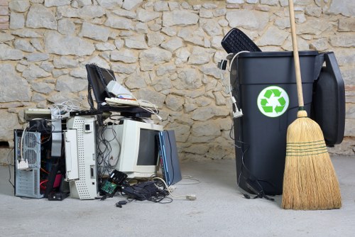 A collection of discarded electronic waste and office equipment, including desktop computers, monitors, keyboards, and cables, piled together on a concrete surface adjacent to a stone wall. To the right, a large black recycling bin with a green recycling symbol is situated next to a wooden broom leaning against it. The background features a beige, textured stone wall, and the scene appears to be outdoors or in a semi-covered area typical of waste collection points. The electronic waste is not organized and appears to be in the process of disposal or collection, aligning with rubbish removal services in Osterley or the surrounding area.