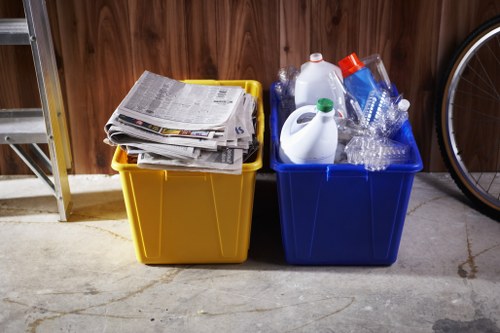 Two plastic bins placed side by side on a concrete floor inside a storage area or garage, with one bin yellow containing a stack of newspapers and magazines on top, and the other bin blue filled with empty transparent and colored plastic bottles, some with caps and labels. The background includes a wooden wall and part of a metal shelving unit, with a bicycle wheel visible to the right. The lighting appears natural or from indoor sources, highlighting the textures of the paper, plastics, and concretes, all contributing to a scene related to waste collection and disposal managed by Commercial Waste Removal Osterley in the Osterley area. The image emphasizes the segregation of recyclable waste materials in preparation for rubbish removal services.
