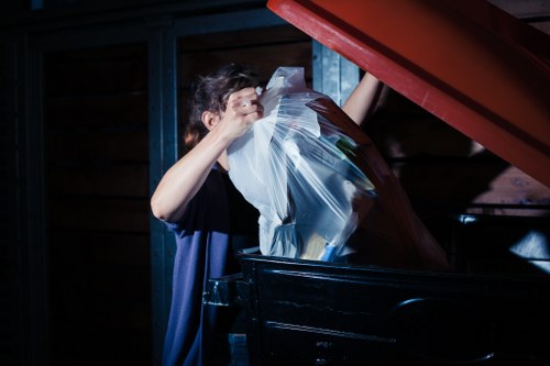A person positioned at a dark-coloured waste collection area, filling a transparent plastic bag with various types of rubbish, including paper and lightweight materials. The individual is wearing a dark shirt and has short, curly hair, with their face partially obscured as they focus on loading the waste. The setting appears to be outdoors or in a storage shed, with wooden walls and blue structural supports visible in the background. The waste container or skip is black with a red lid lifted open to facilitate disposal. The scene is illuminated by natural or ambient lighting, highlighting the textures and colors of the waste and surrounding area. This image relates to commercial waste removal services in Osterley, demonstrating the process of disposing of mixed refuse efficiently and safely, as provided by Commercial Waste Removal Osterley, ensuring waste is managed appropriately for local businesses and properties in the area.