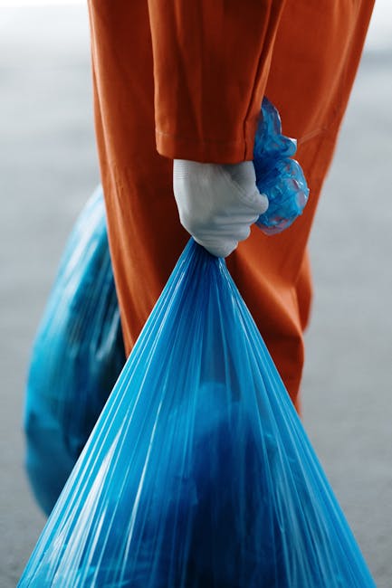A person standing on a concrete floor is holding two large blue plastic rubbish bags, one in each hand. The individual is dressed in an orange coverall and wearing white footwear, with their hands cov